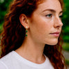 Close-up of a woman wearing gold and green earrings with a blurred green background